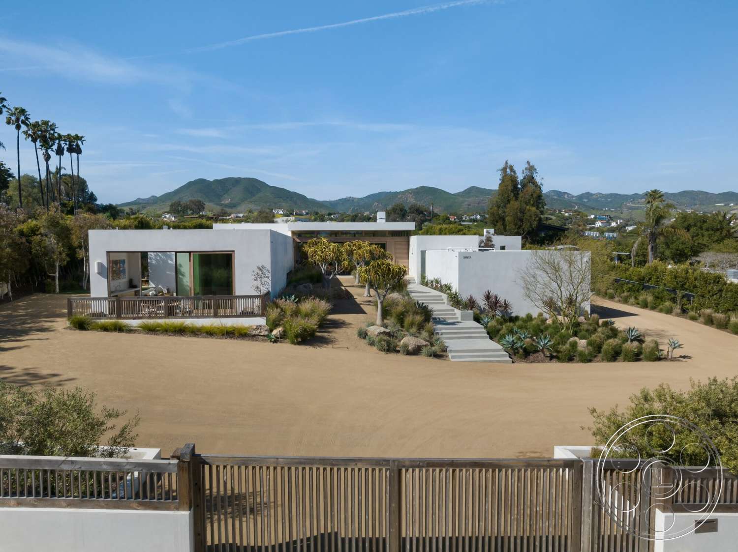 Modern 529 - exterior, concrete steps, paved walkway, open outdoor space, concrete driveway, minimalist architectural style, wooden deck railing, large panoramic windows, landscaped garden with succulents, surrounding mountain view, flat-roofed modern home, palm trees in the background, desert-themed garden