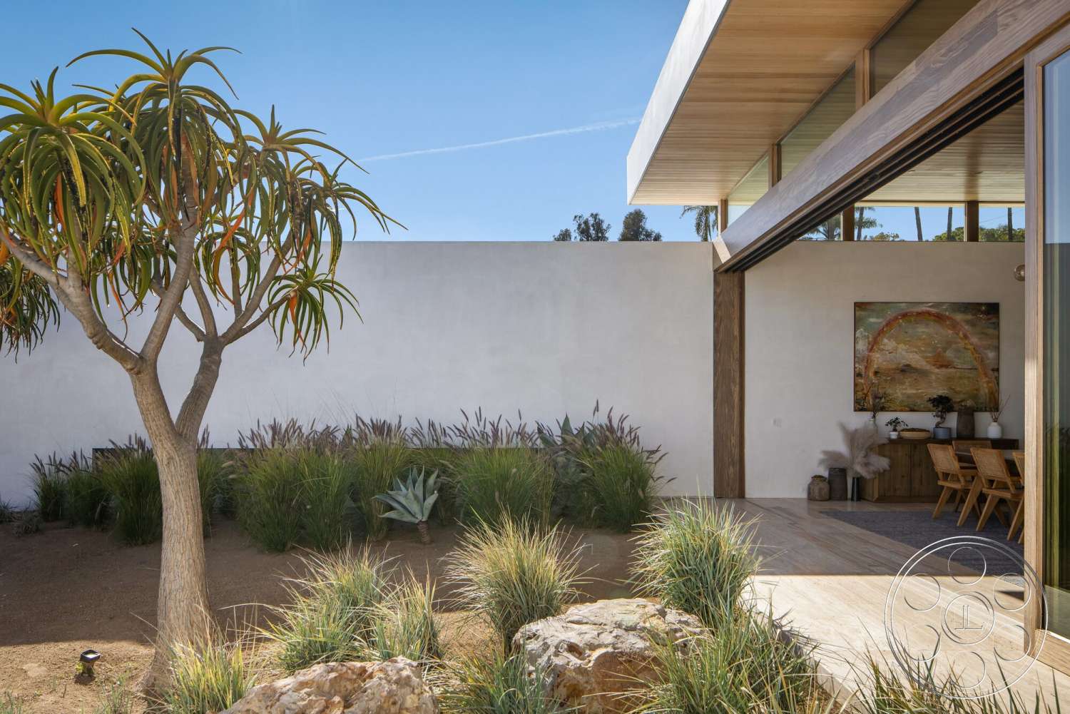 Modern 529 - exterior, white stucco walls, large glass sliding doors, natural wood accents, framed artwork, clear blue sky, desert landscaping, modern garden design, wood beam roof overhang, minimalist architectural style, concrete patio floor, planted grass area, decorative stone boulders, dining area adjacent to garden, tropical tree