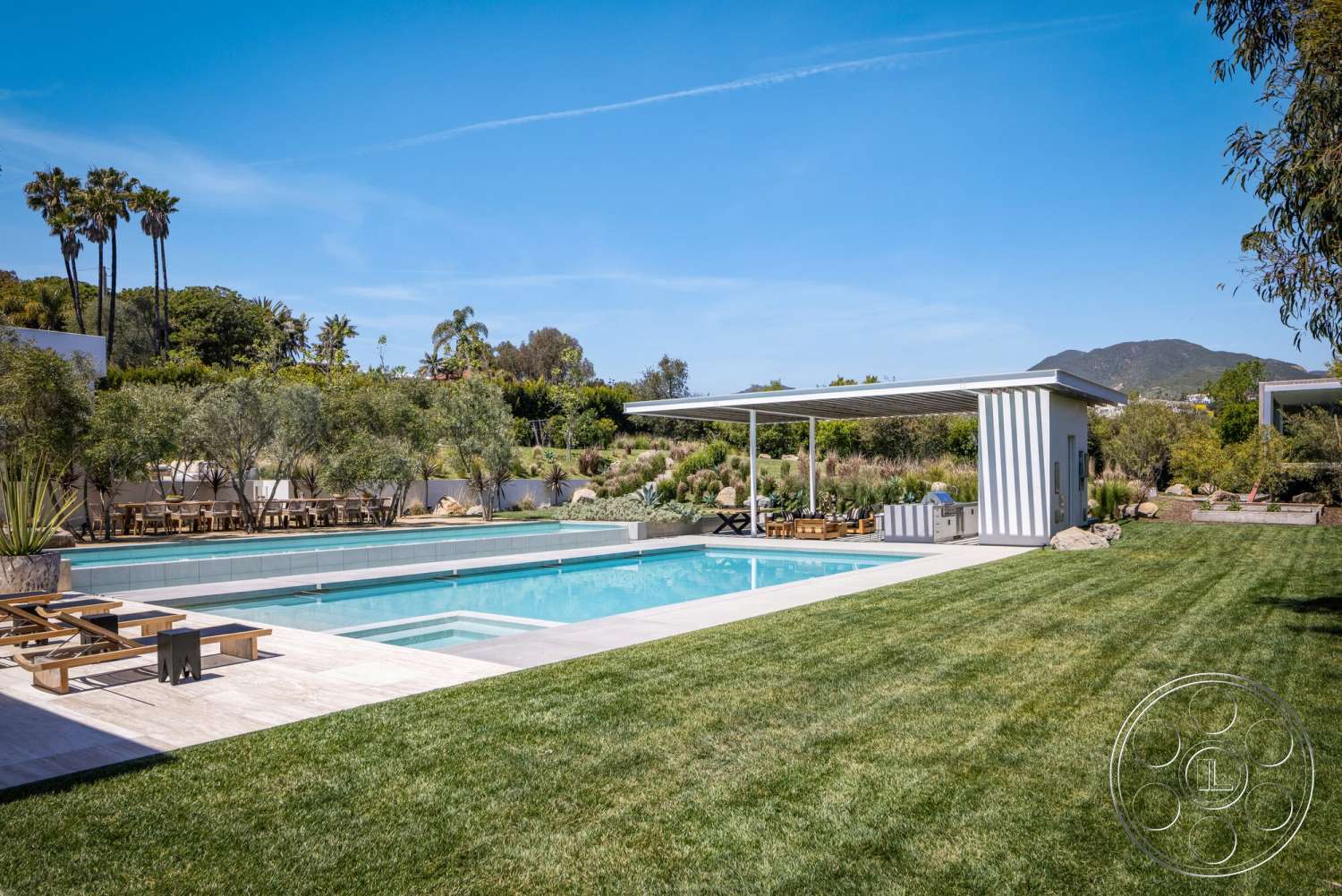 Modern 529 - exterior, palm trees, mountain view, green grass lawn, rectangular pool, wooden lounge chairs, stone pool deck, landscaped garden area, concrete patio border, pergola with slatted roof, modern outdoor kitchen, planter boxes with shrubs