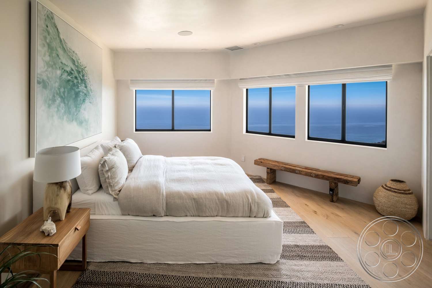 Modern 528 - textured area rug, plaster wall treatment, floor-to-ceiling windows with ocean view, minimalist bedroom design, light hardwood bedroom floors, decorative woven basket, wooden bench with rustic finish, modern bedside lamp with natural wood base, abstract wall art featuring blue and green tones, white bed linens with layered textures