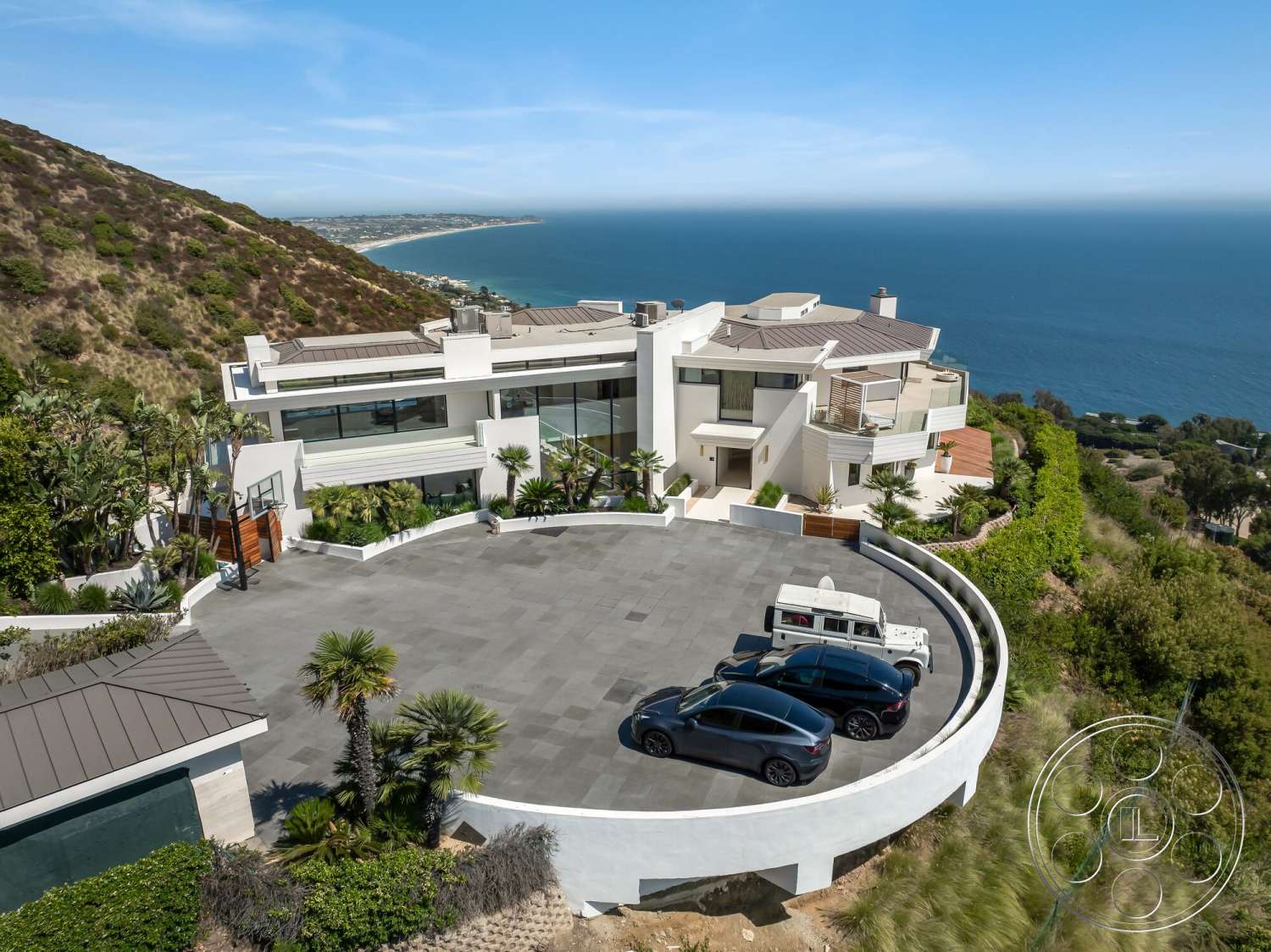 Modern 528 - exterior, landscaped garden, white stucco walls, floor-to-ceiling windows, circular driveway, modern architectural style, mountain backdrop, palm tree landscaping, ocean view setting, gray stone pavers, metal roof shingles, designed outdoor space