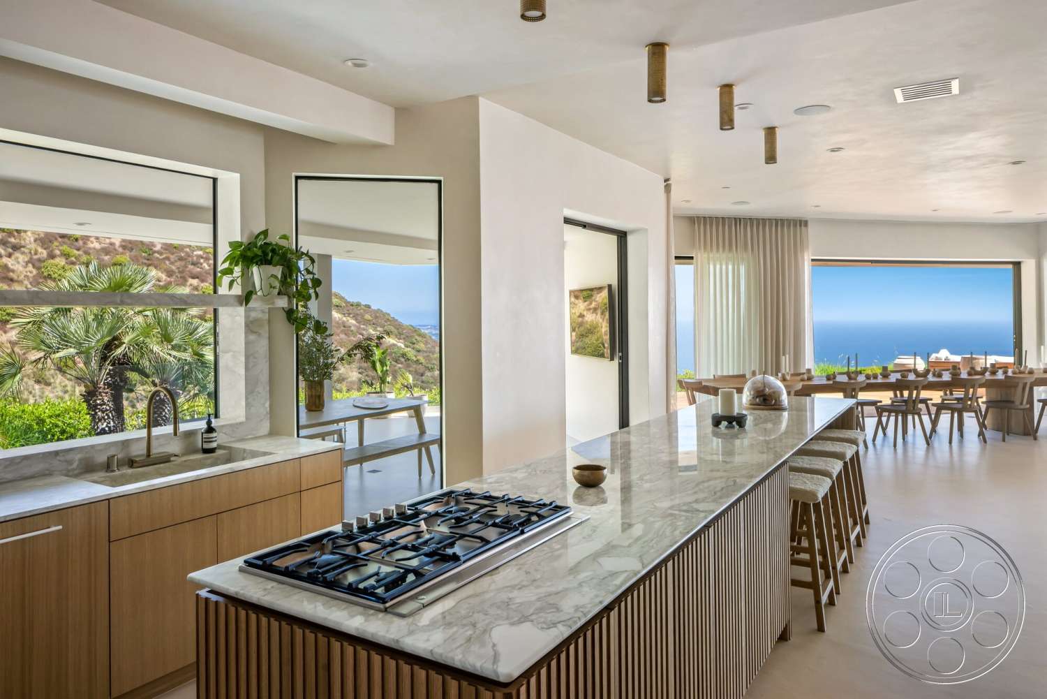 Modern 528 - natural light, open floor plan, woven bar stools, wood cabinetry, floor-to-ceiling windows, large sliding glass doors, white painted walls, smooth plaster walls, minimalist lighting fixtures, stainless steel gas cooktop, marble kitchen island countertops, dining area with ocean view, elevated hillside setting