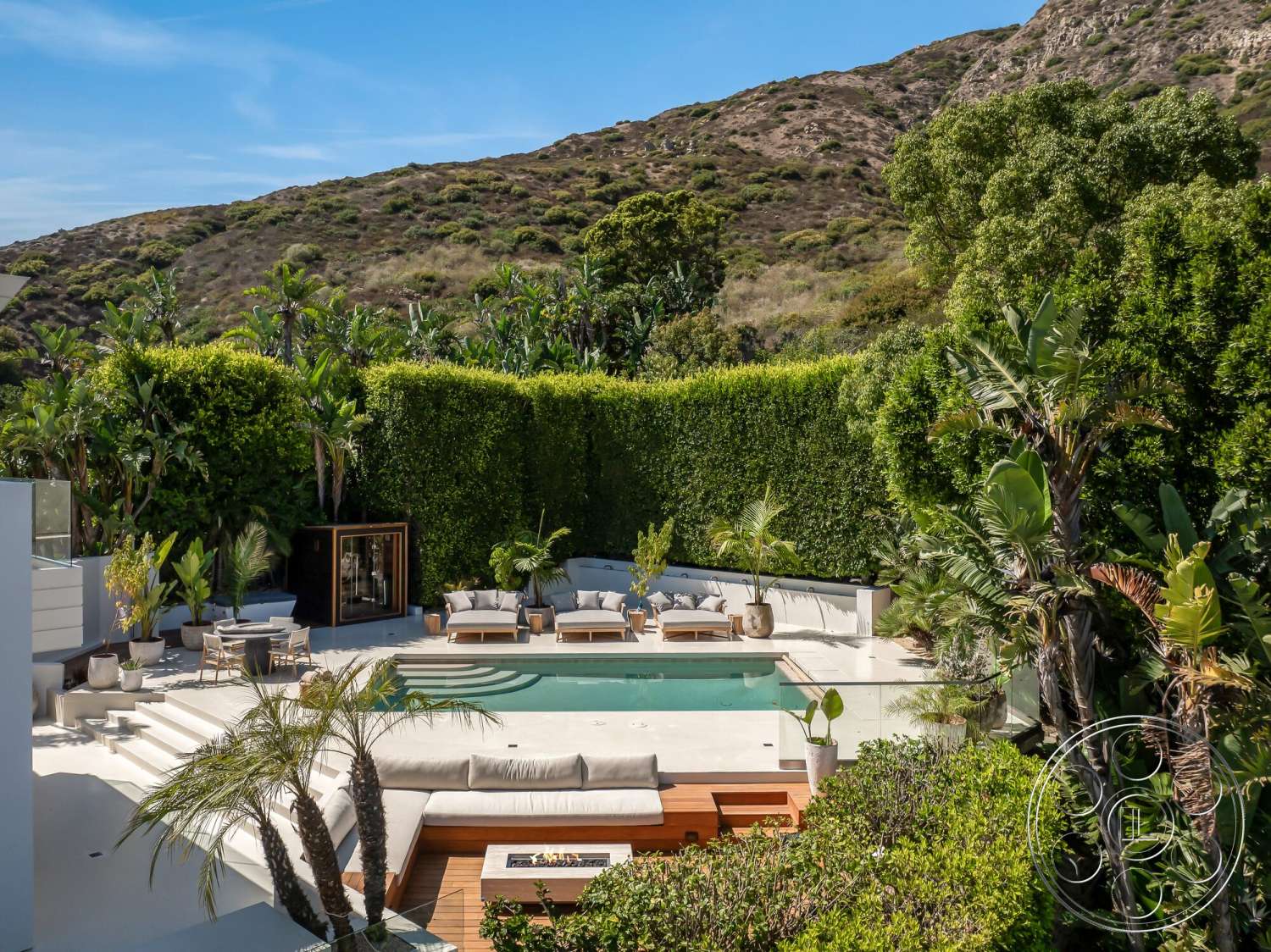 Modern 528 - exterior, potted plants, tropical landscaping, open-air living space, lush green hedges, concrete pool deck, rectangular pool, natural stone steps, stone fire pit, modern patio furniture, planted garden beds, mountain view setting, wood deck seating area, wood lounge chairs, glass fencing around pool