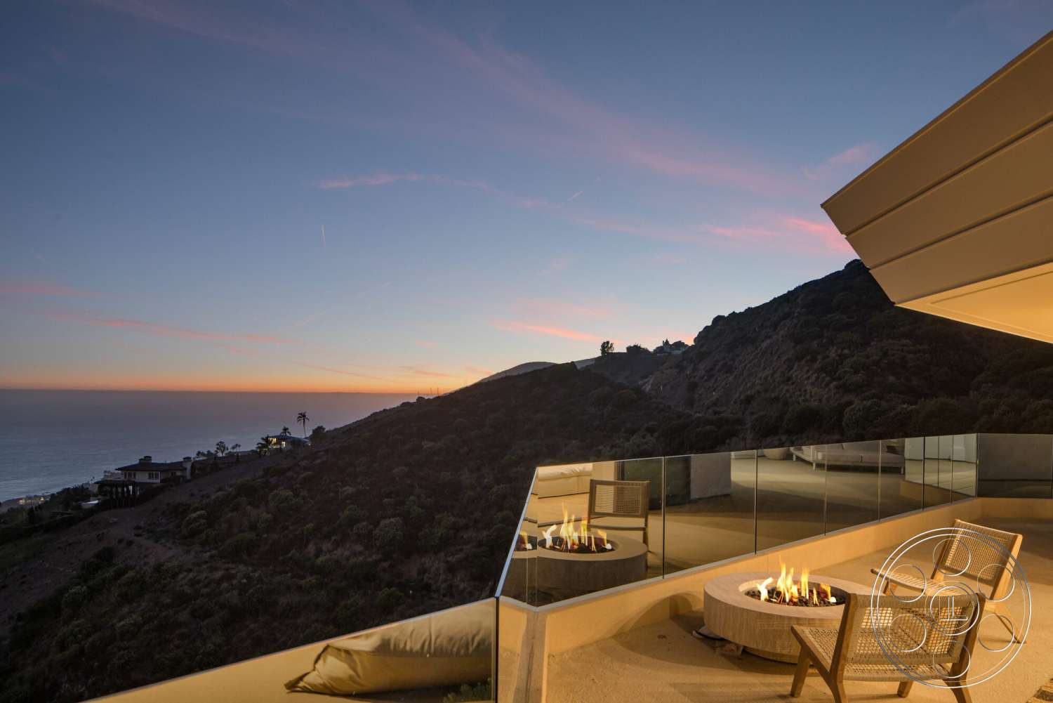Modern 528 - exterior, modern architectural style, ocean view, concrete patio flooring, large overhang roof, mountain hillside setting, glass railing around patio, fire pit with circular concrete surround, outdoor seating area with wooden chairs, sunset sky with pink and orange hues
