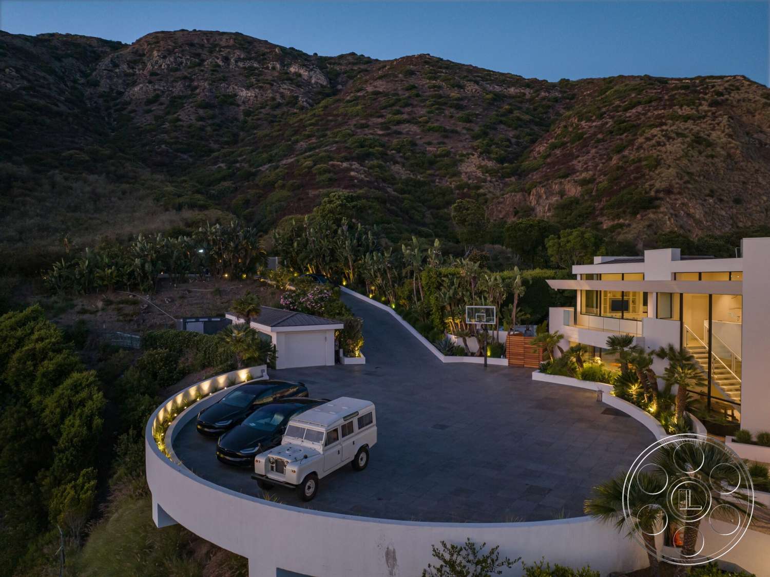 Modern 528 - exterior, flat roof, floor-to-ceiling windows, curved driveway, architectural lighting, landscaped garden area, surrounding mountain view, modern house design, outdoor basketball hoop, paved driveway made of stone, urban-rural blend setting, multiple vehicle parking space, palms landscaping