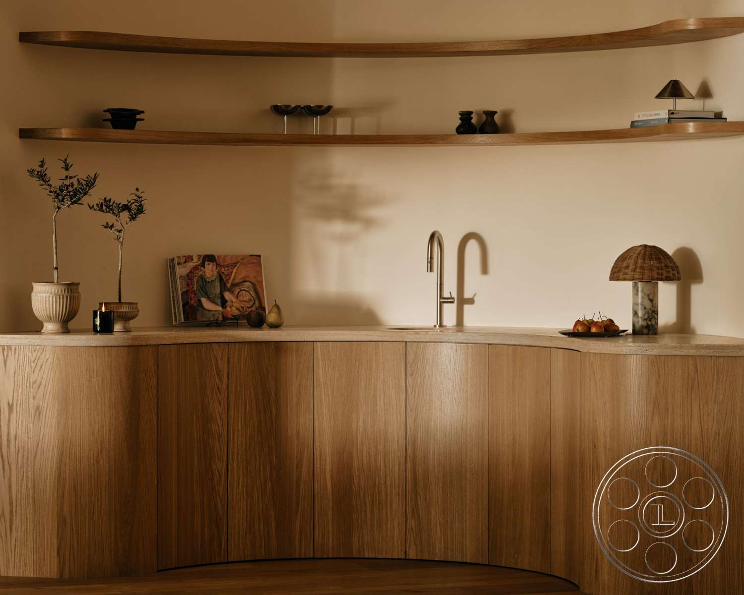 Modern 531 - neutral color palette, soft ambient lighting, light wood countertop, textured plaster wall, open wood shelving, brass kitchen faucet, glass lamp base, curved wood cabinetry, ceramic plant pot, stone fruit bowl, decorative art book