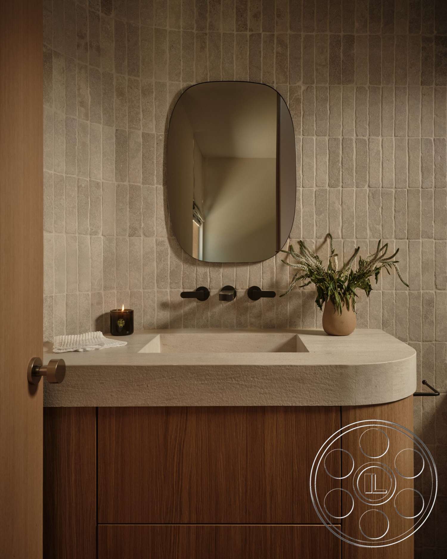 Modern 531 - minimalist decor, wood cabinetry, small potted plant, oval mirror, black wall-mounted faucets, textured stone bathroom walls, rounded concrete sink countertop