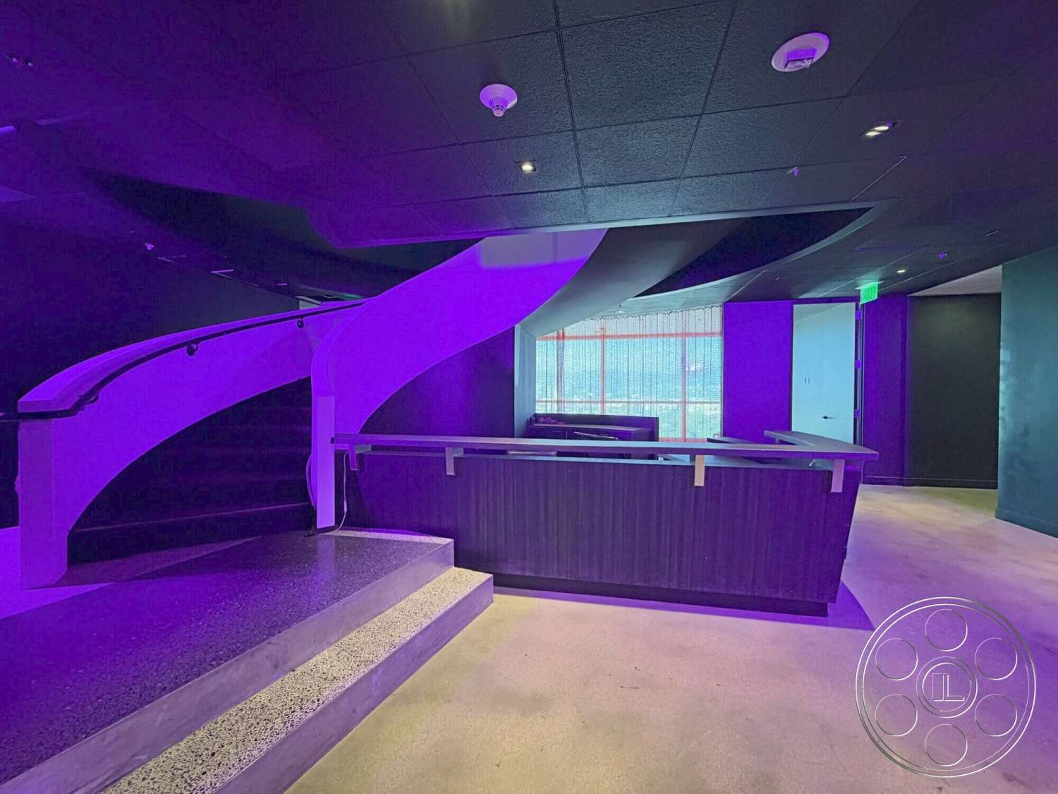 Wilshire Penthouse - open floor plan, polished concrete flooring, textured wall finish, floor-to-ceiling windows, curved staircase, modern reception desk, black ceiling tiles, exposed metal railings, multicolored lighting accents