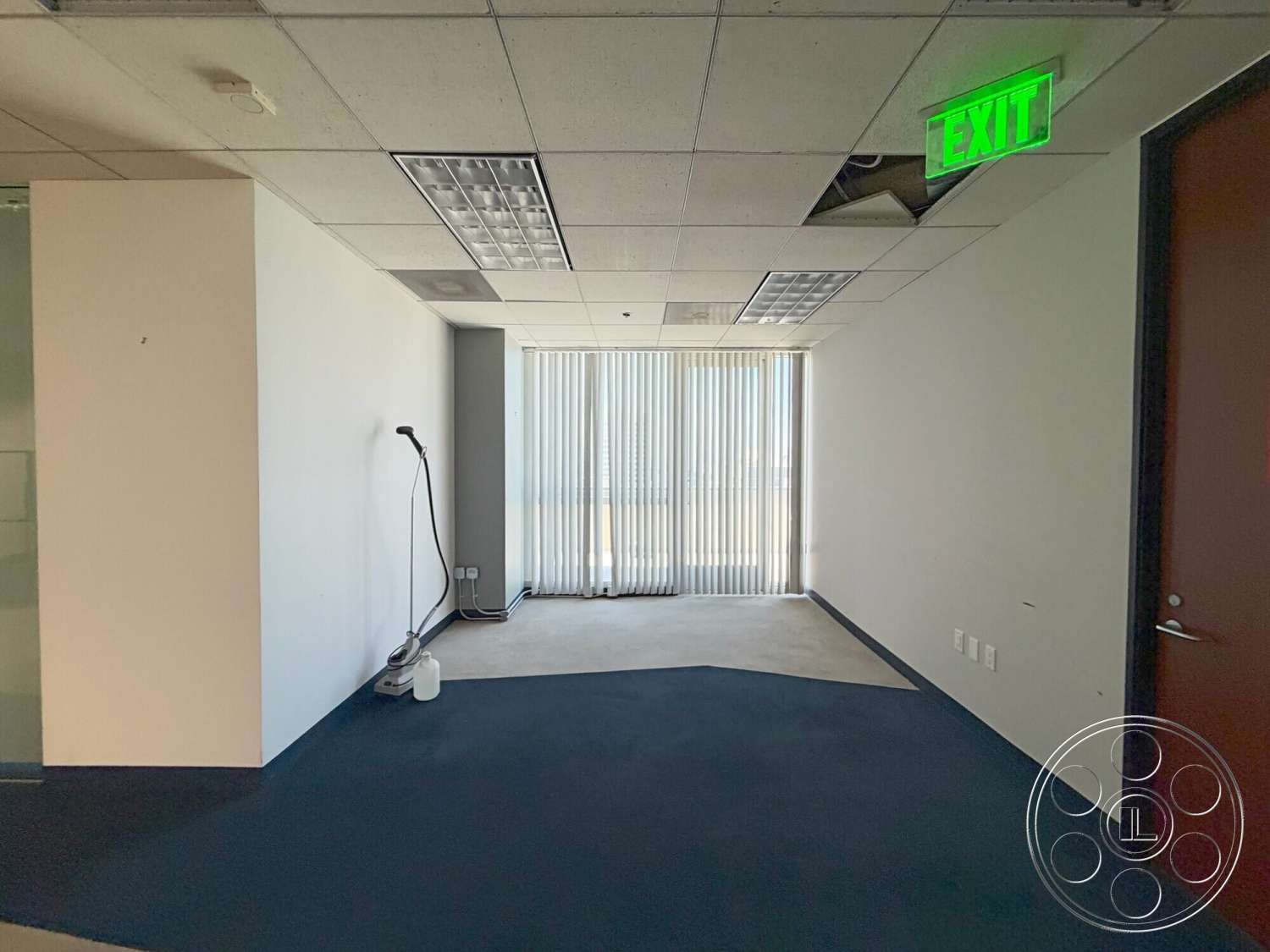 Wilshire Penthouse - carpet flooring, window with vertical blinds, exit sign, drop ceiling tiles, glass partition wall, fluorescent ceiling lights, drywall walls, blue carpet floor, doorway with wooden door