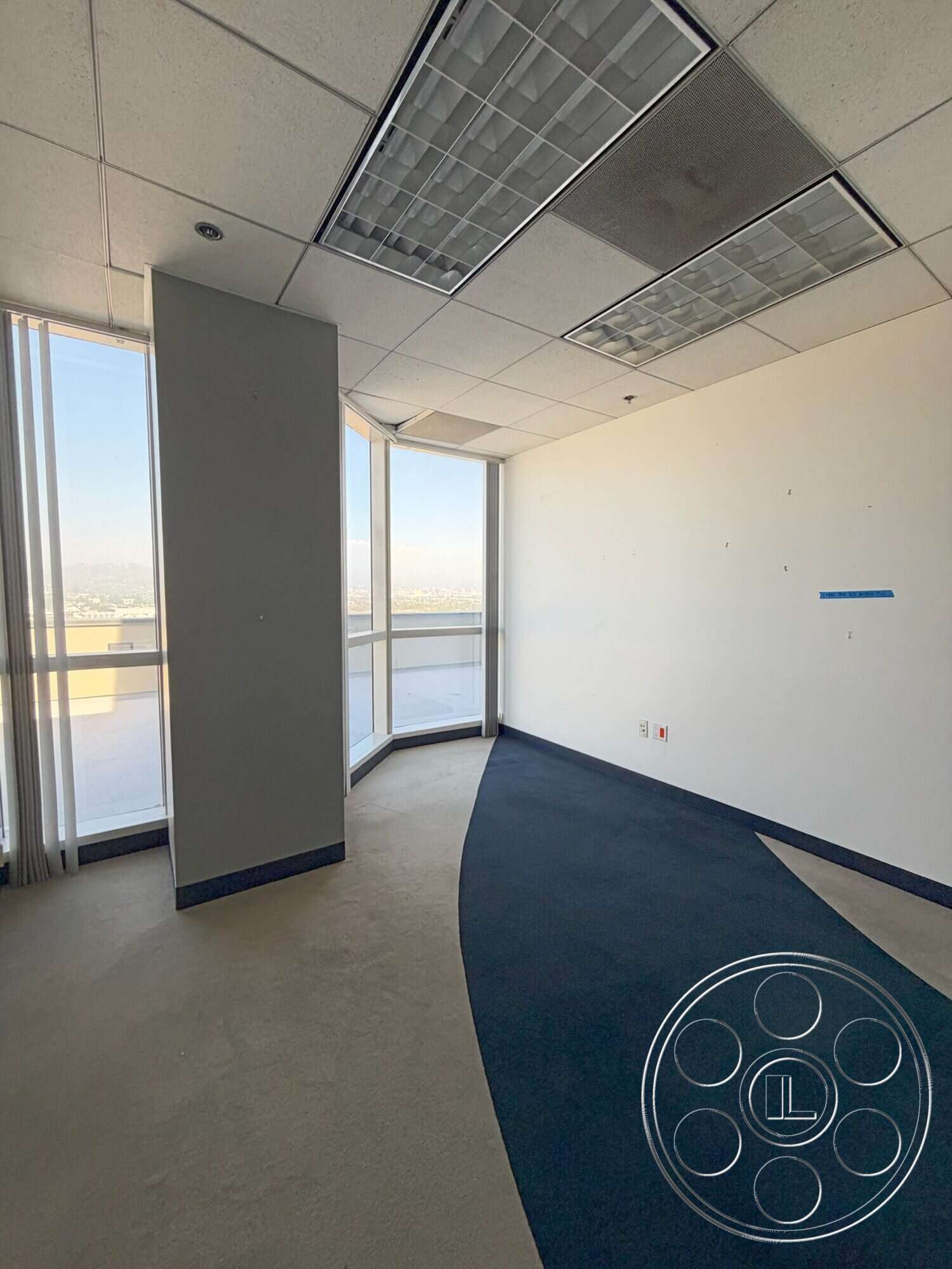 Wilshire Penthouse - carpet flooring, neutral color scheme, white painted walls, interior office space, large windows with city view, drop ceiling with fluorescent lighting, blue carpet floor with curved design