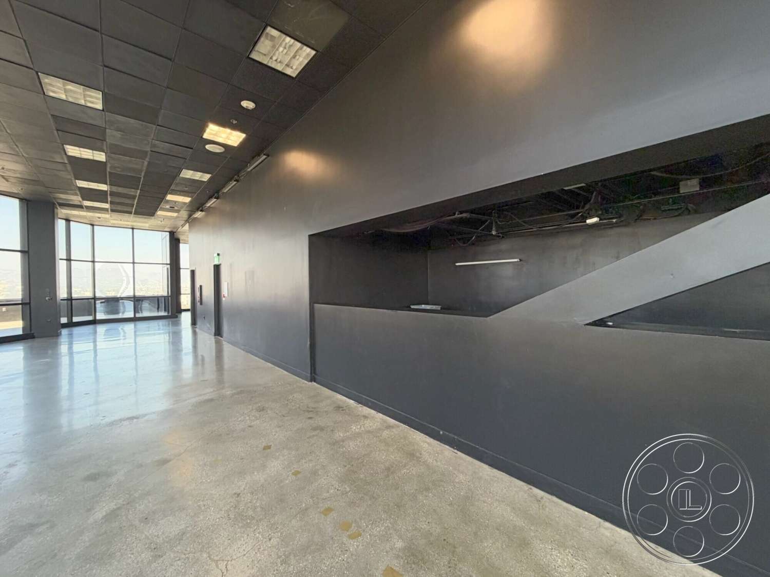 Wilshire Penthouse - open floor plan, polished concrete flooring, minimalist design elements, interior space, large floor-to-ceiling windows, black painted walls, urban high-rise setting, exposed electrical wiring, modern industrial aesthetic, drop ceiling with square grid tiles, linear wall cutout for service area