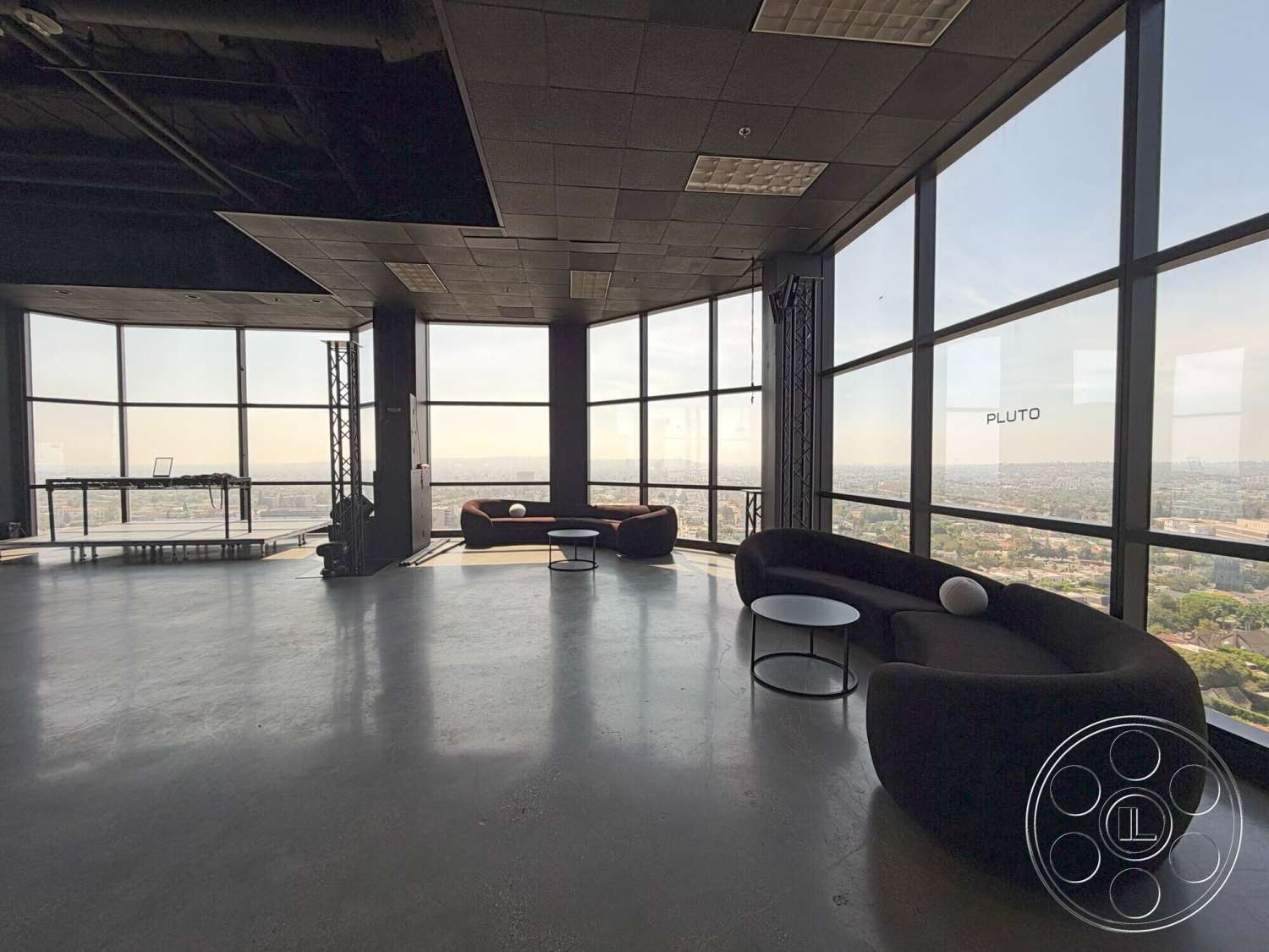 Wilshire Penthouse - minimalist decor, round coffee table, open floor plan, modern furniture, polished concrete flooring, floor-to-ceiling windows, urban skyline view, curved sofa design, black acoustic ceiling tiles, metal frame structure, exposed ceiling ductwork