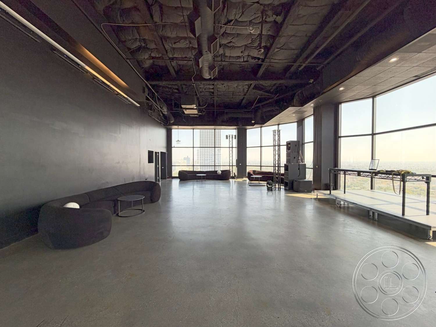 Wilshire Penthouse - interior space, modern coffee table, floor-to-ceiling windows, city skyline view, black painted walls, open floor layout, industrial ceiling design, exposed concrete floors, black sectional sofa, steel frame stage