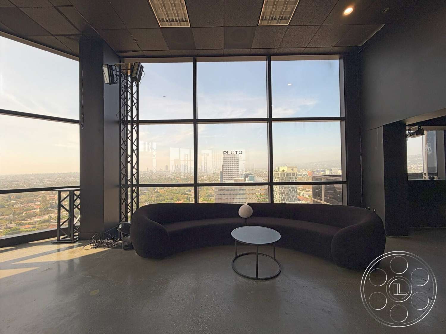 Wilshire Penthouse - concrete flooring, modern coffee table, floor-to-ceiling windows, dark wall finishes, industrial-style lighting fixtures, exposed structural framework, open urban skyline view, black fabric curved sofa