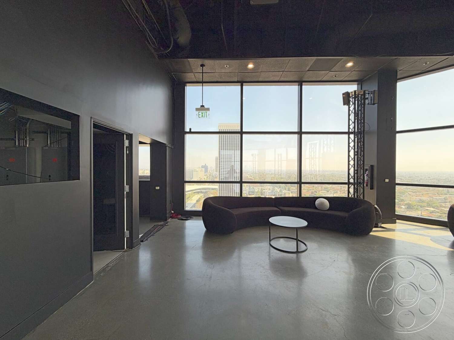 Wilshire Penthouse - round coffee table, interior space, floor-to-ceiling windows, urban skyline view, contemporary interior design, curved sectional sofa, black painted walls, exposed concrete flooring, industrial ceiling with visible ductwork