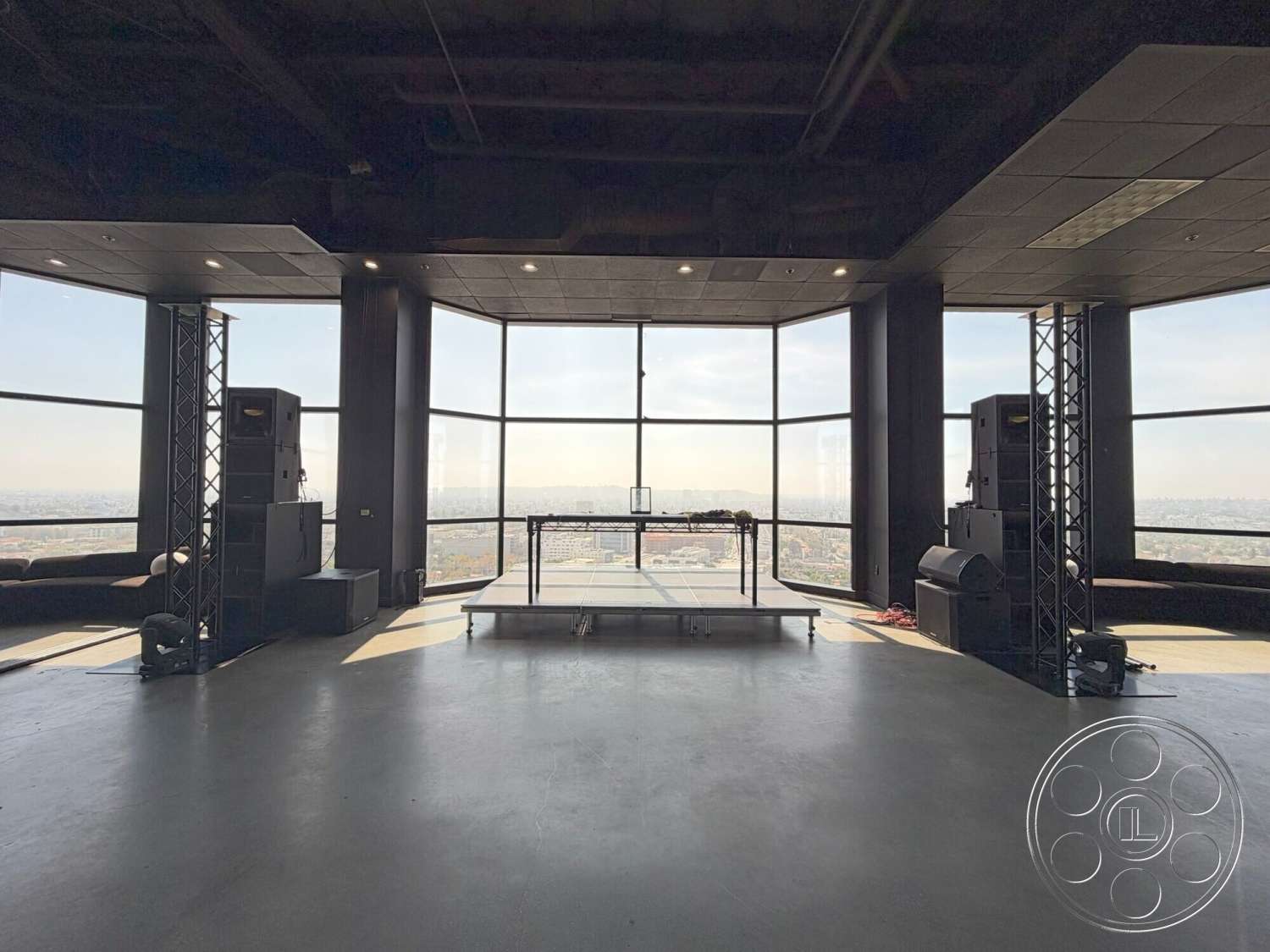 Wilshire Penthouse - concrete flooring, open space layout, panoramic city skyline view, black steel support columns, exposed steel structural elements, glass wall windows, flush LED ceiling lights, industrial-style design, sound system equipment, black ceiling tiles, low-profile stage, recessed lighting features