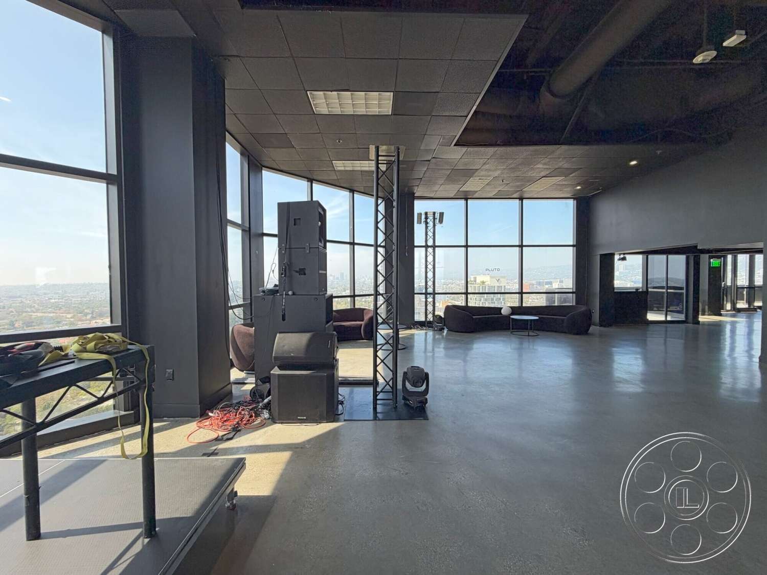 Wilshire Penthouse - open floor plan, polished concrete flooring, minimalist furniture, floor-to-ceiling windows, exposed structural elements, industrial-style lighting, black acoustic ceiling tiles, performance stage area, large space with urban skyline view, seating area with modern couches, electronic sound equipment