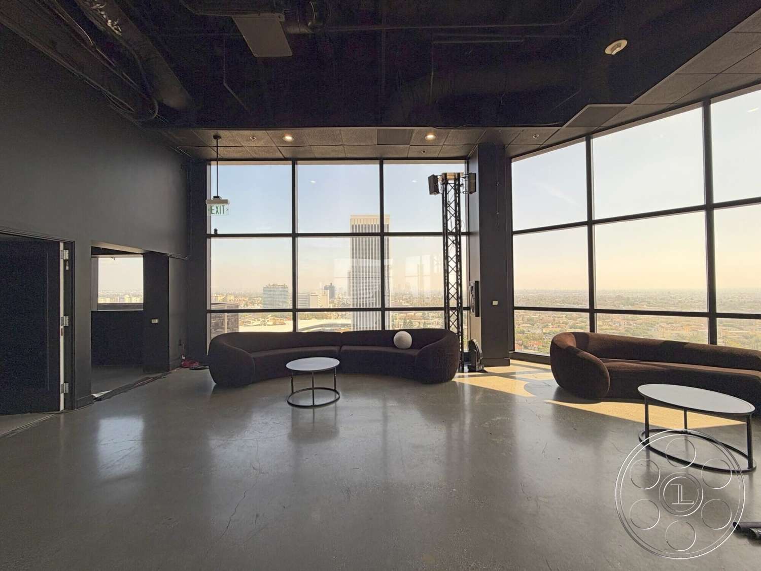 Wilshire Penthouse - round coffee table, open floor plan, concrete flooring, floor-to-ceiling windows, modern architectural style, curved sectional sofa, black painted walls, industrial-style lighting, exterior city skyline view