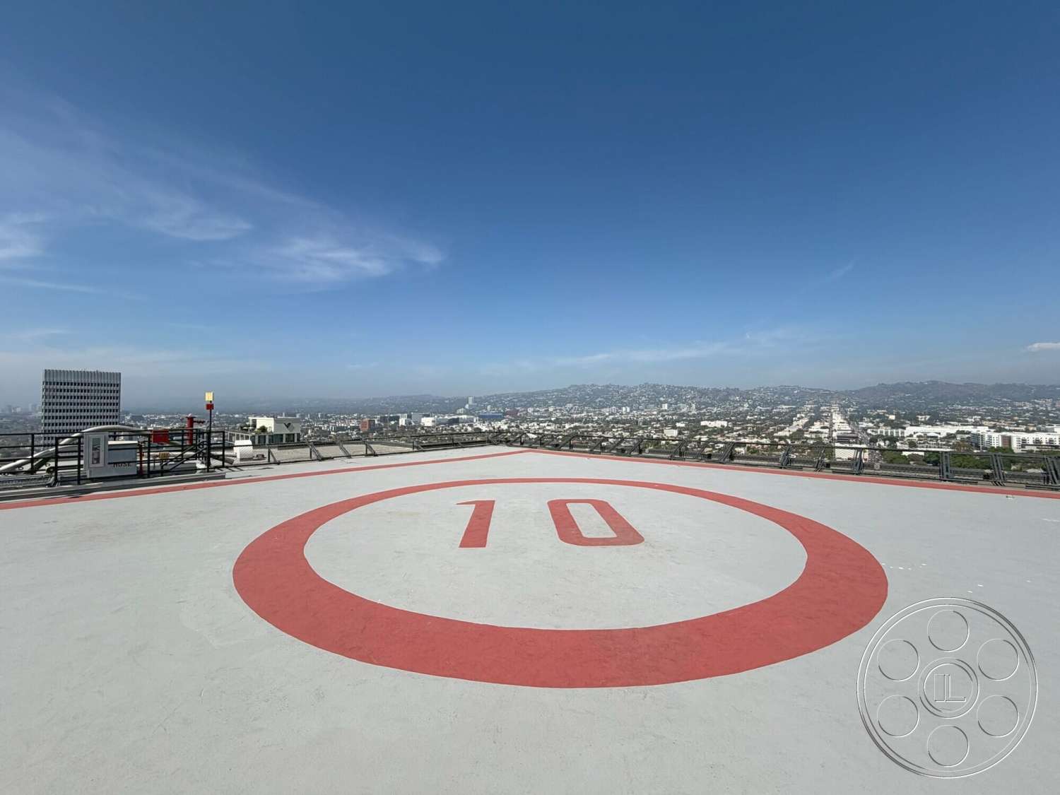 Wilshire Penthouse - exterior, helicopter pad, urban skyline view, metal railing perimeter, concrete helicopter landing area, red painted circular markings, open rooftop setting