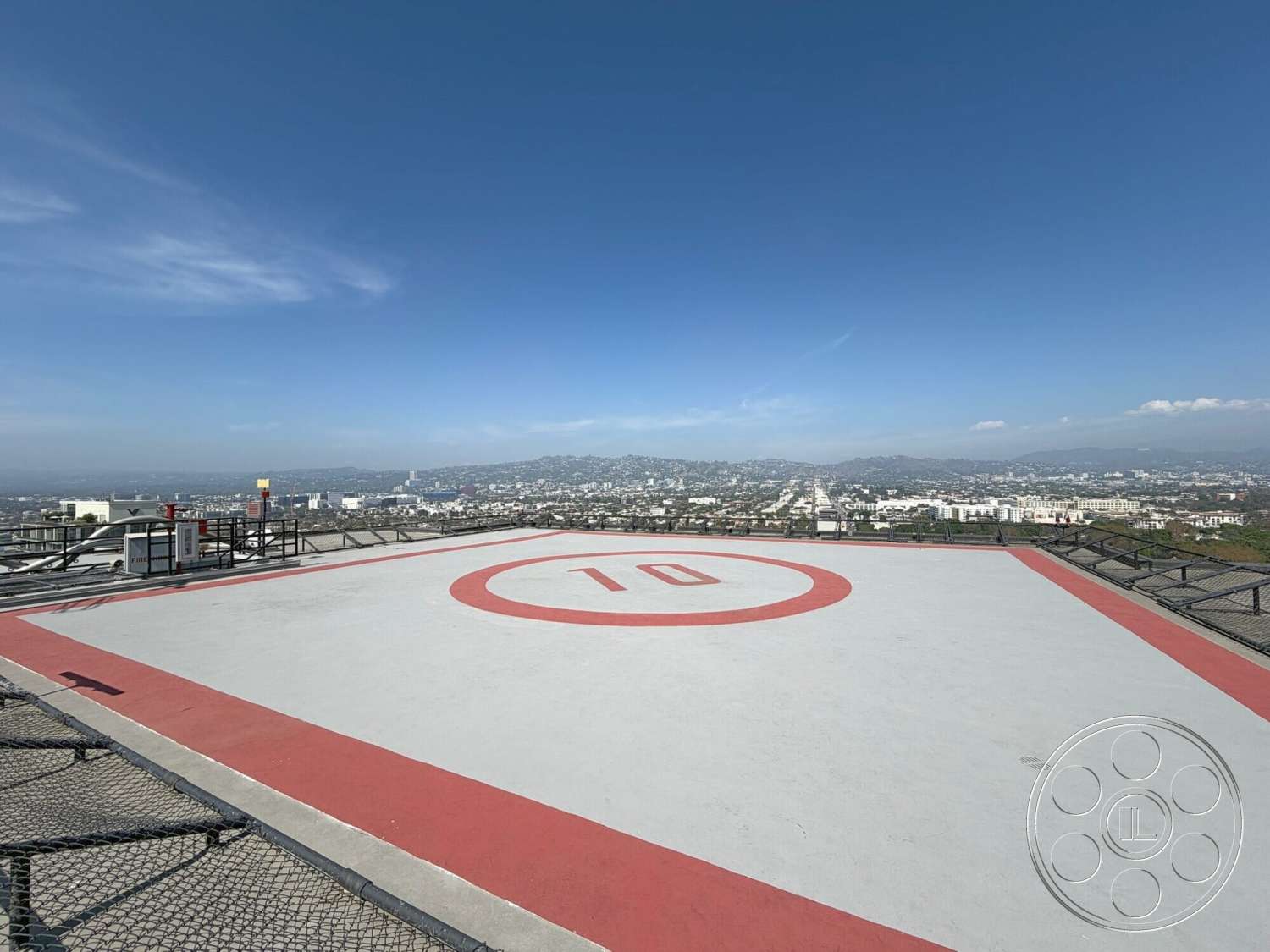 Wilshire Penthouse - exterior, urban skyline view, clear blue sky, mountainous backdrop, concrete helipad surface, metal safety railing, white painted concrete with red markings, rectangular helipad shape