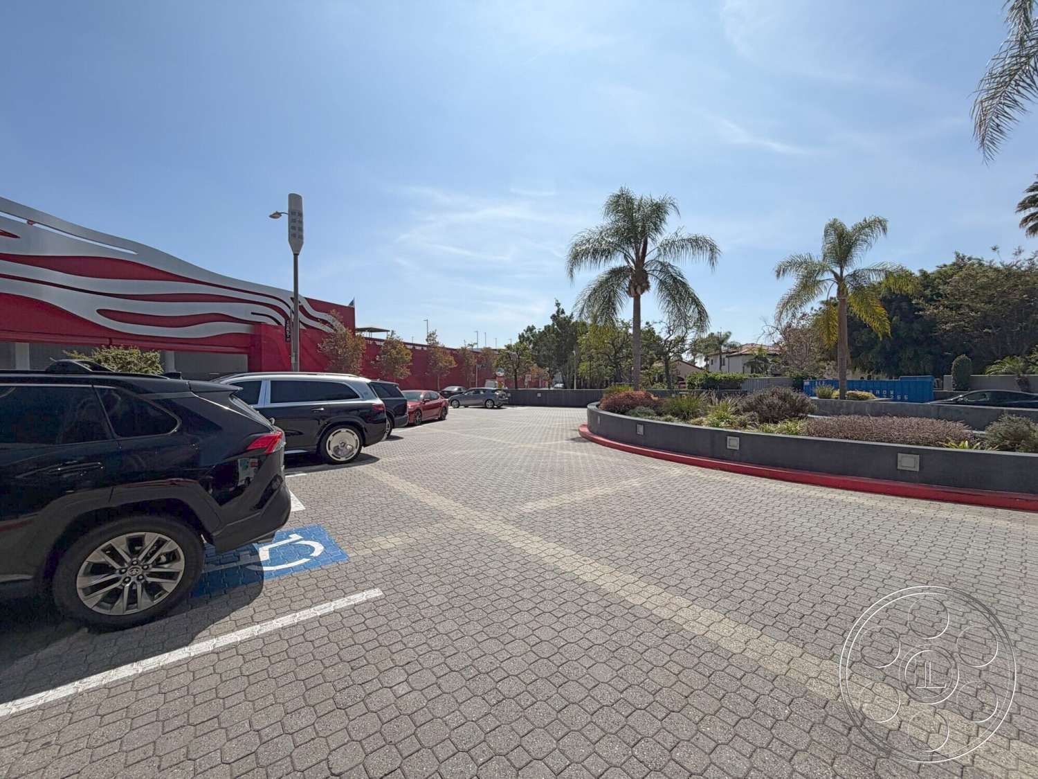 Wilshire Penthouse - palm trees, blue sky, paved parking lot, outdoor environment, accessible parking space, brick paver driveway, modern architectural design, red painted building exterior, curved landscape planter, commercial property exterior