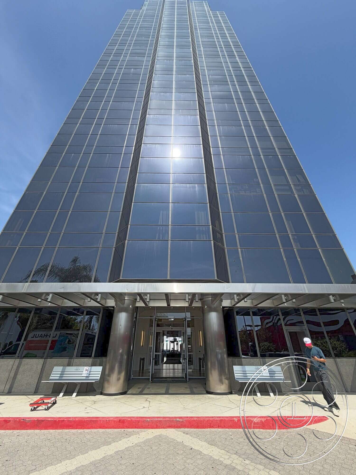 Wilshire Penthouse - exterior, modern architectural style, steel columns, landscaped entrance area, concrete building base, reflective glass panels, glass curtain wall façade, polished stone entrance floor, aluminum framing, cantilevered roof overhang, large automatic entrance doors, symmetrical building shape