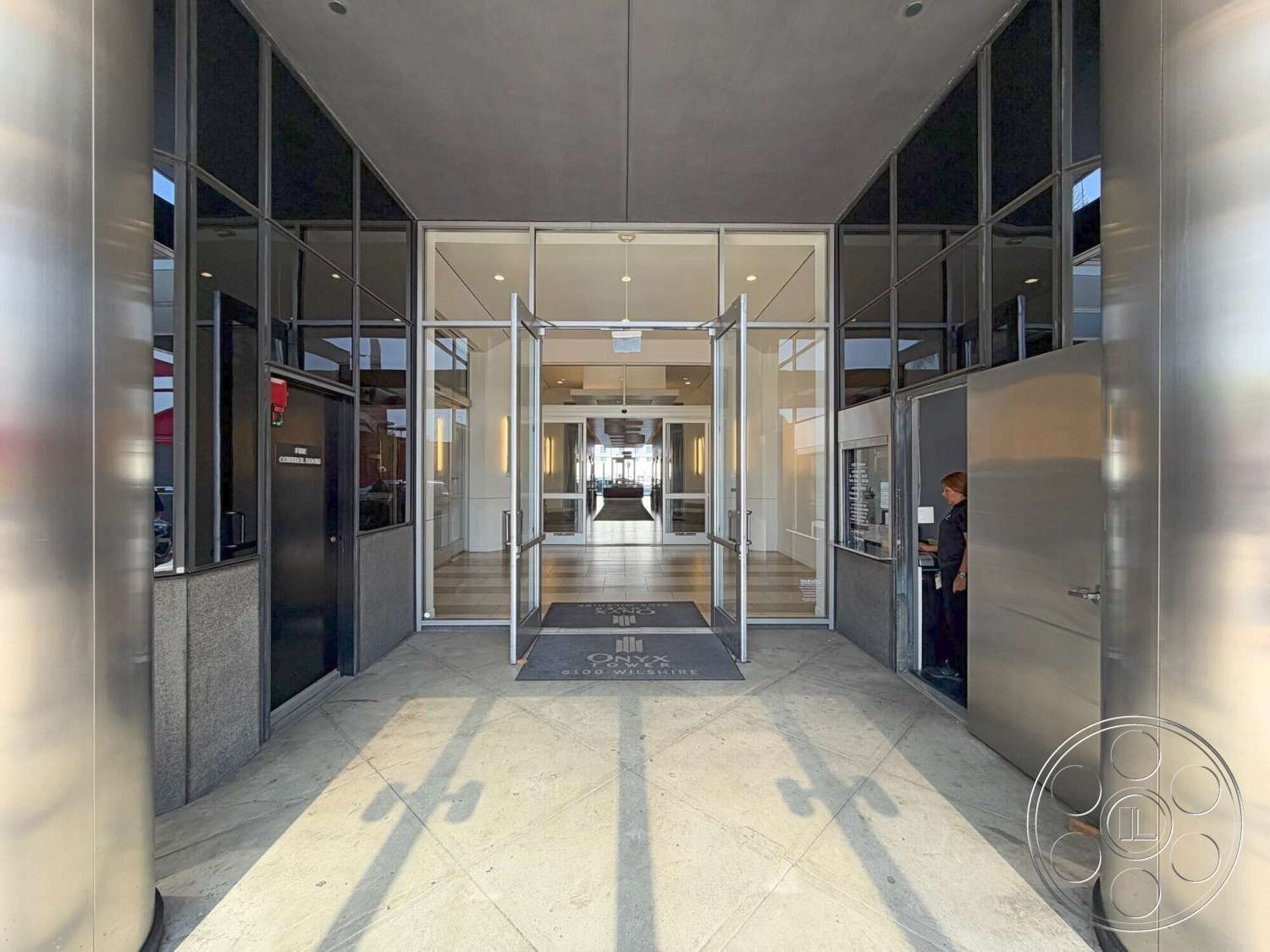 Wilshire Penthouse - exterior, polished concrete floor, modern architectural style, floor-to-ceiling glass windows, glass entrance doors, granite wall cladding, linear ceiling lighting, metal door frames, open lobby entrance, slate welcome mat, metal paneling details, broad concrete walkway, vertical support columns