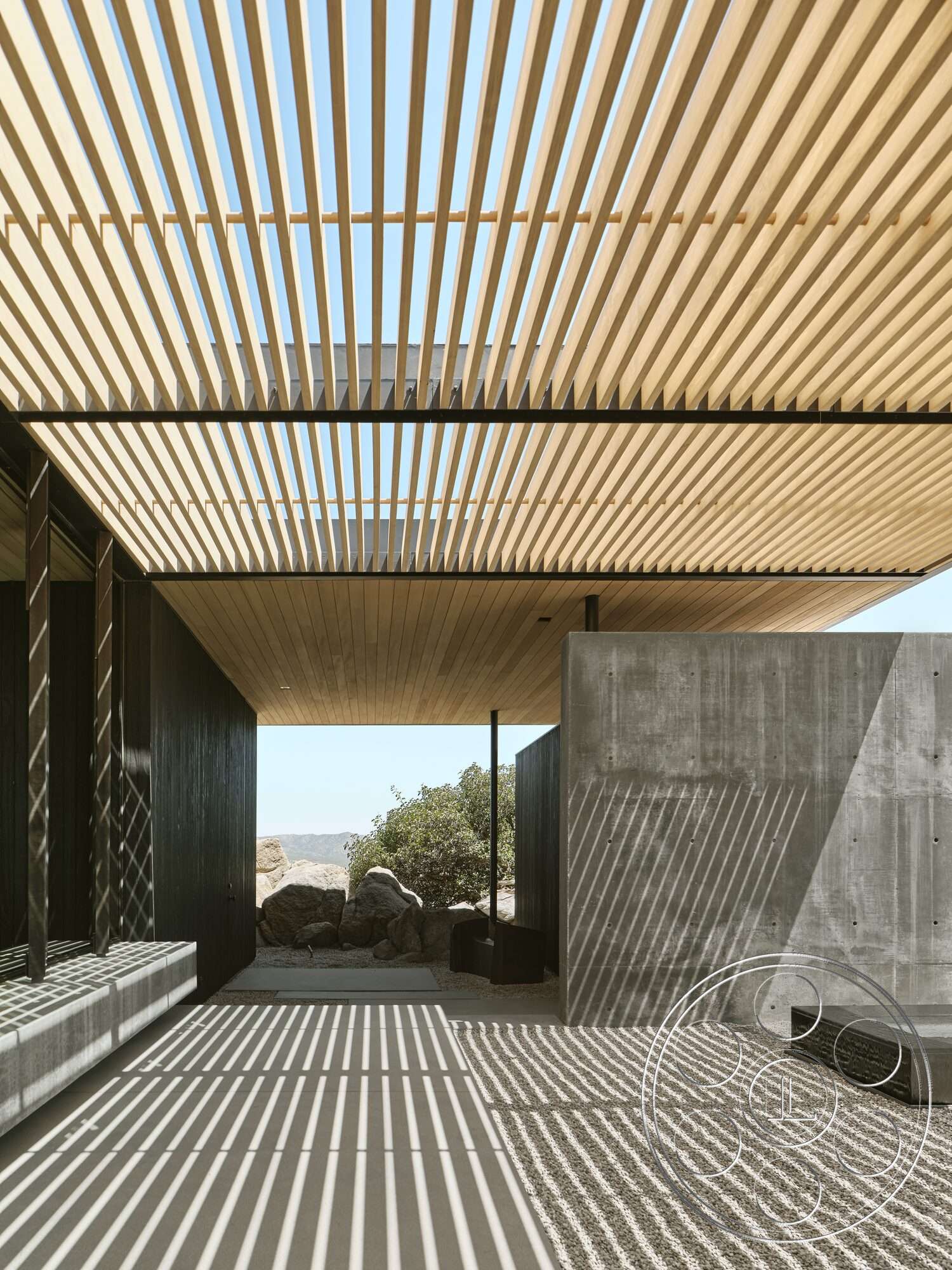 Palm Springs 110 - exterior, concrete wall, horizontal wood paneling, gravel patio flooring, minimalist architectural design, stone landscaping features, desert landscape setting, rectangular outdoor space, wood slat roof overhang, exposed concrete textures, black steel structural elements, natural light filtering through slats