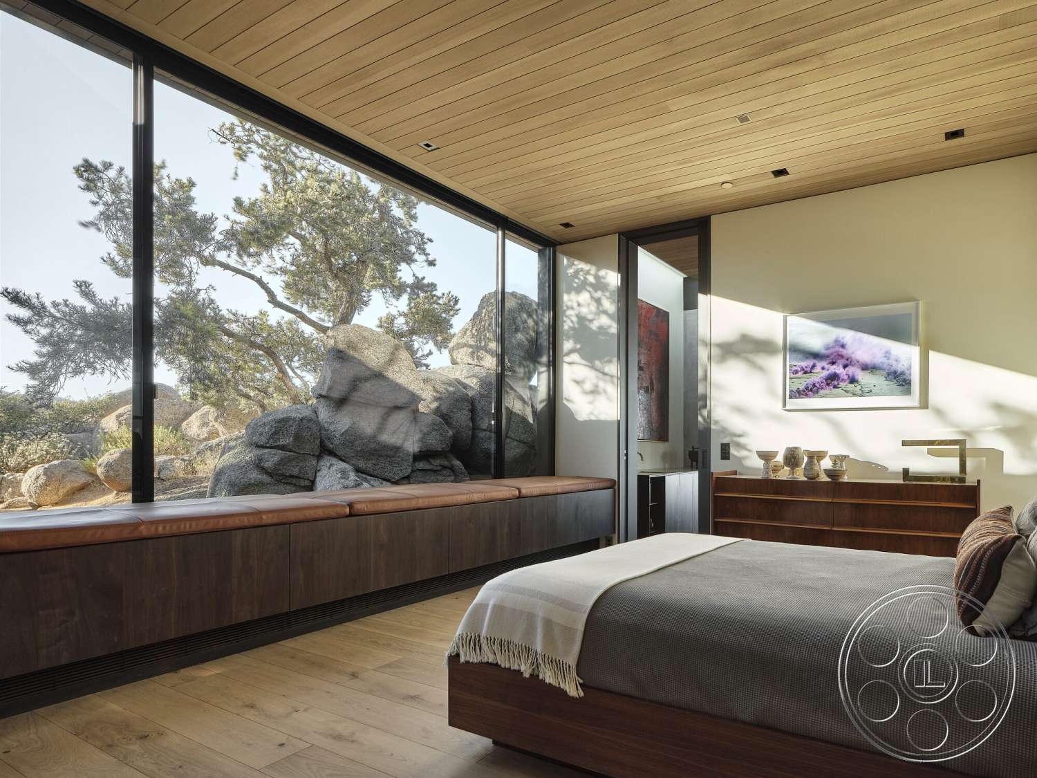 Palm Springs 110 - dark wood cabinetry, stone accent wall, floor-to-ceiling windows, artwork on wall, natural light exposure, modern minimalistic design, contemporary bedroom, light hardwood bedroom floors, wood ceiling planks, wood window seat, rugged rock formation view, textured blanket on bed