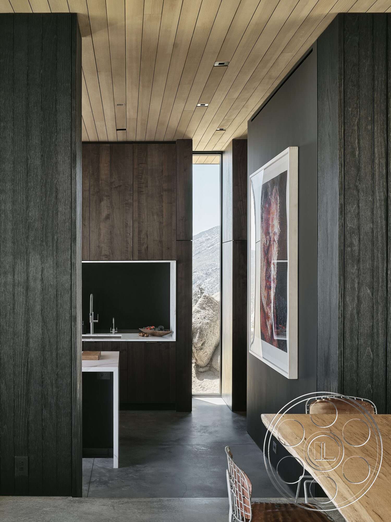 Palm Springs 110 - floor-to-ceiling windows, exposed concrete floor, light wood ceiling panels, modern dining room table, dark wood paneling kitchen cabinetry, black painted kitchen wall