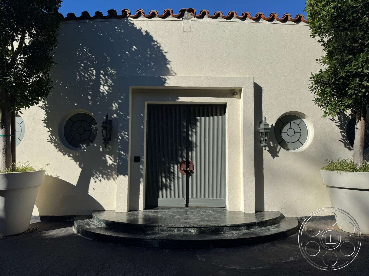 The Hearst House - exterior, landscaped planters, terracotta roof tiles, stucco walls, decorative tile details, exposed light fixtures, wooden front door, circular windows, green garden shrubs, rounded front steps, marble entry steps, glass door panels