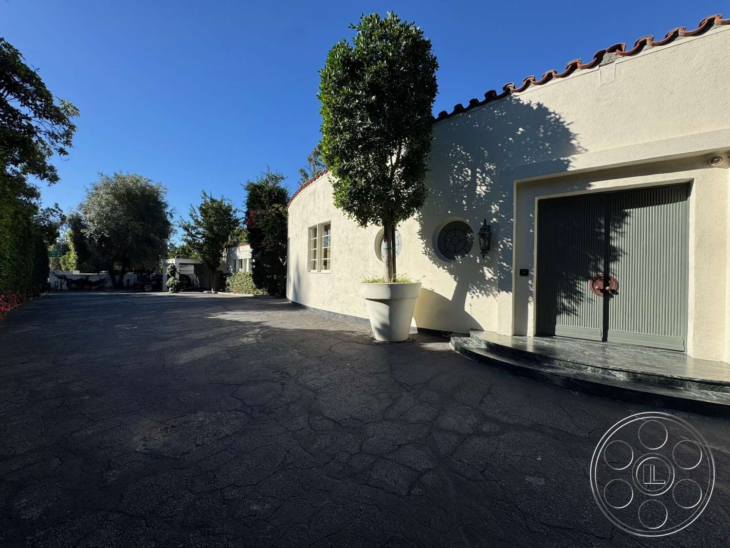 The Hearst House - exterior, modern landscape design, curved driveway, red tile roof, stucco walls, circular window, large potted plant, textured concrete driveway, shadow play from trees, circular planter