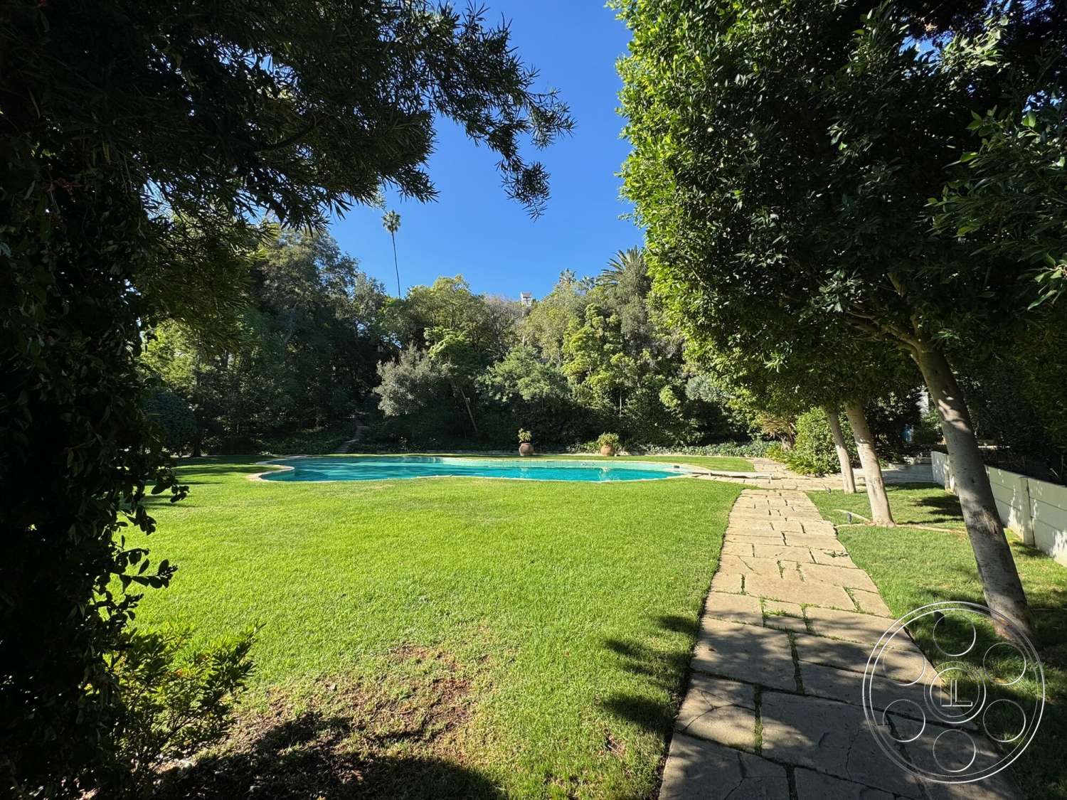 The Hearst House - exterior, natural landscaping, lush green lawn, surrounded by trees, flagstone pathway, oval swimming pool, concrete pool border, sunny backyard setting