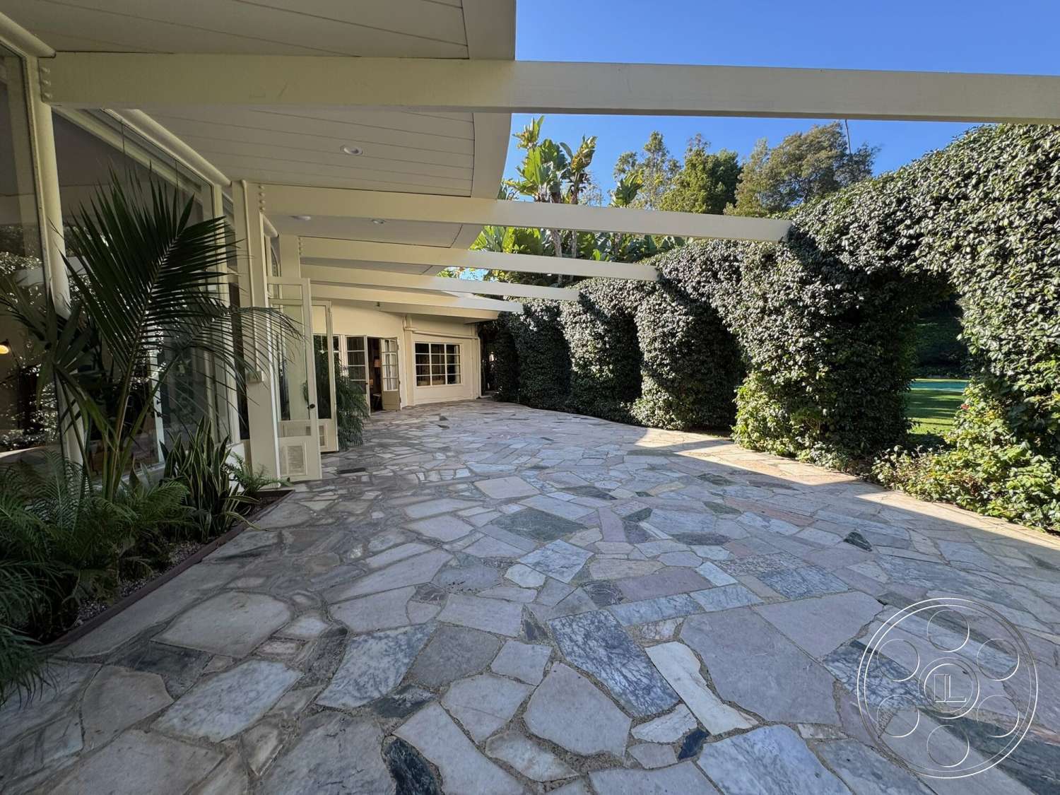 The Hearst House - exterior, floor-to-ceiling windows, tropical landscaping, open-air terrace, flat roof design, lush green foliage, natural stone patio flooring, covered outdoor area, rounded hedge wall, light wooden support beams