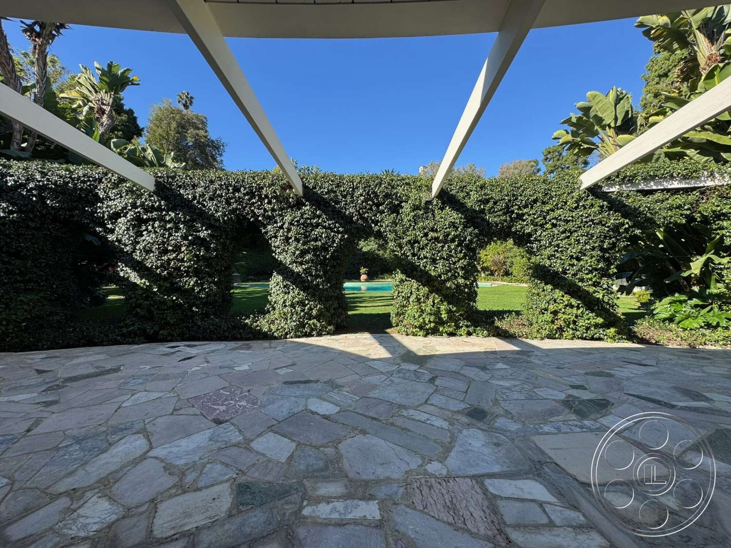 The Hearst House - exterior, natural stone pavers, landscaped garden view, lawn area, stone patio flooring, lush greenery and palm trees, arched trellis with climbing plants, modern structural beams, shaded outdoor seating space