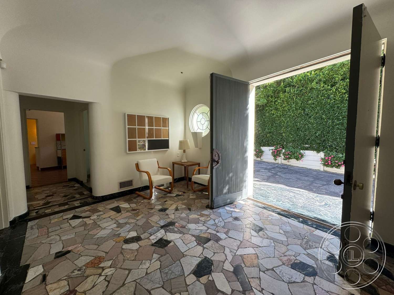 The Hearst House - minimalist furniture, wooden side table, arched doorway, white painted walls, natural light from entryway, large double doors, multicolored stone flooring, granite border accent in entryway, decorative circular window, comfortable accent chairs, lush green exterior landscaping