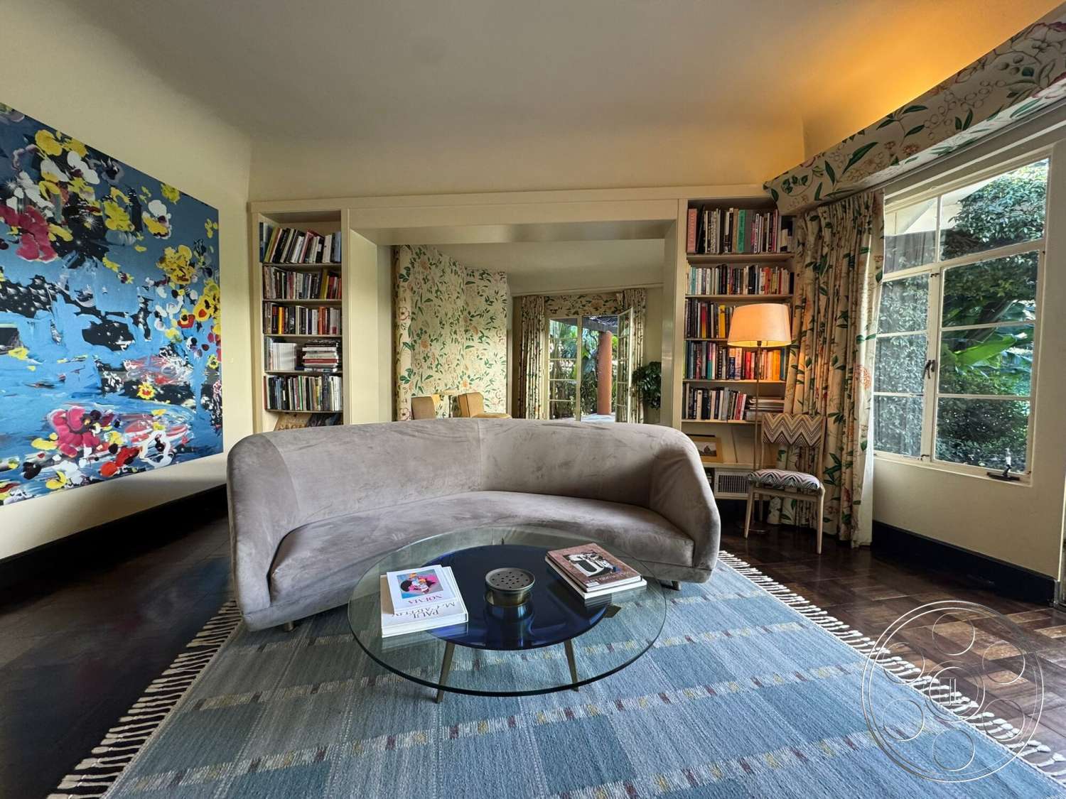The Hearst House - natural light, hardwood flooring, textured area rug, glass coffee table, decorative plant, floor-to-ceiling windows, built-in bookshelf, open living space, floral patterned curtains, cozy reading nook, modern lighting fixture, neutral painted walls, colorful abstract wall art, plush curved sofa, chevron accent chair