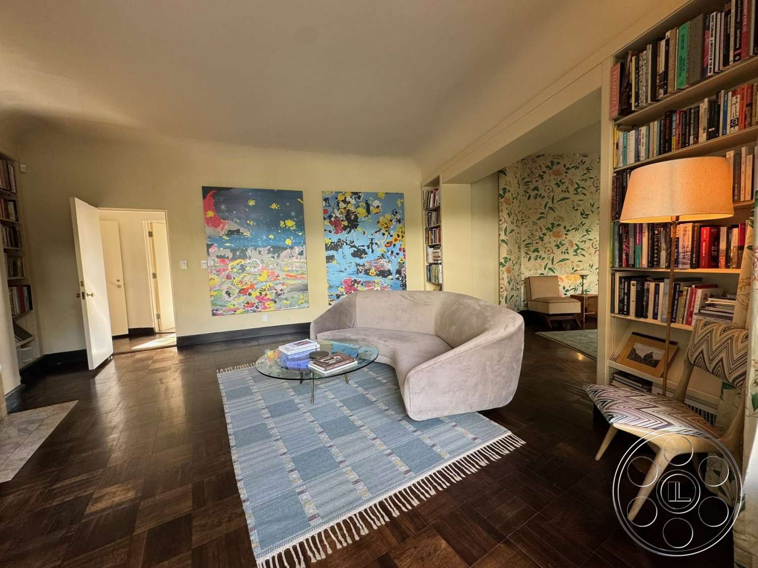 The Hearst House - hardwood flooring, textured wall art, glass coffee table, floral wallpaper, neutral wall color, woven area rug, curved sofa, bookshelf with wooden shelves