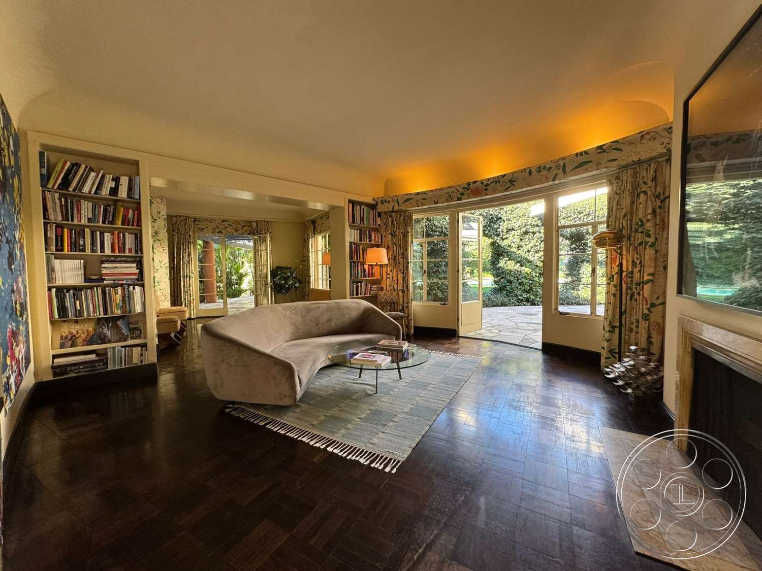 The Hearst House - natural light, open floor plan, glass coffee table, floor-to-ceiling windows, textured ceiling, built-in bookshelf, accent lighting, floral patterned curtains, outdoor garden view, stone fireplace surround, wooden door frames, parquet hardwood living room floors, yellow painted walls, curved contemporary sofa