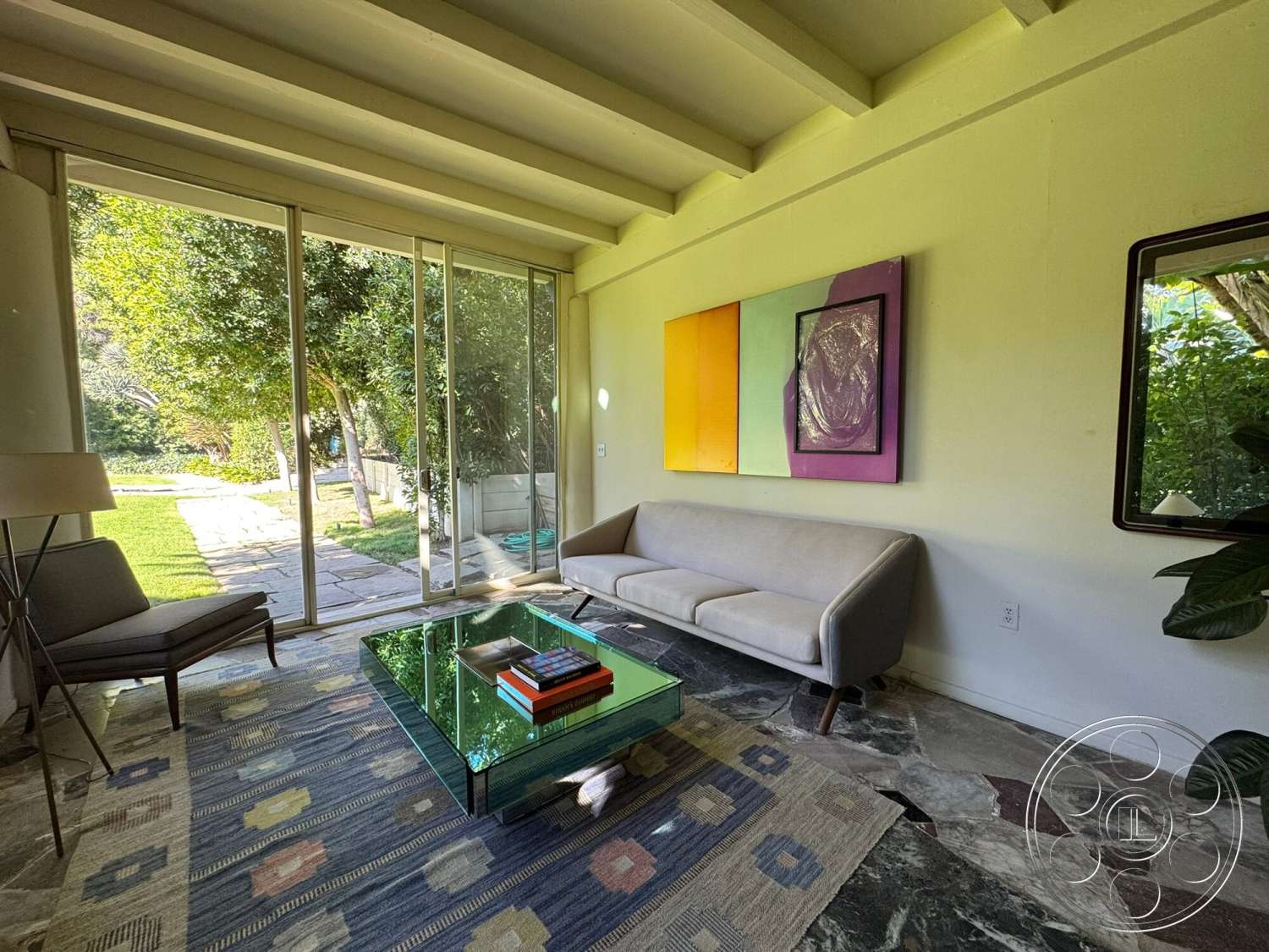 The Hearst House - natural light, glass sliding door, lush garden view, exposed wood ceiling beams, contemporary art wall decor, modern glass coffee table, multicolored patterned area rug, marble living room flooring, neutral-colored upholstered sofa, angled armchair
