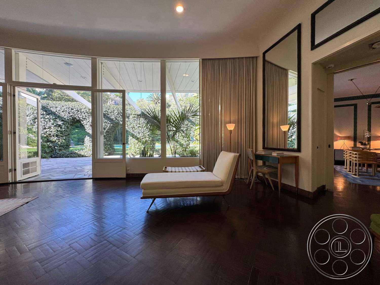 The Hearst House - open floor plan, large glass windows, floor-to-ceiling windows, striped area rug, dark hardwood flooring, soft ambient lighting, arched ceiling, framed mirrors, exposed ceiling beams, contemporary architectural style, potted palm plants, minimalist lounge chair, modern desk with wooden legs