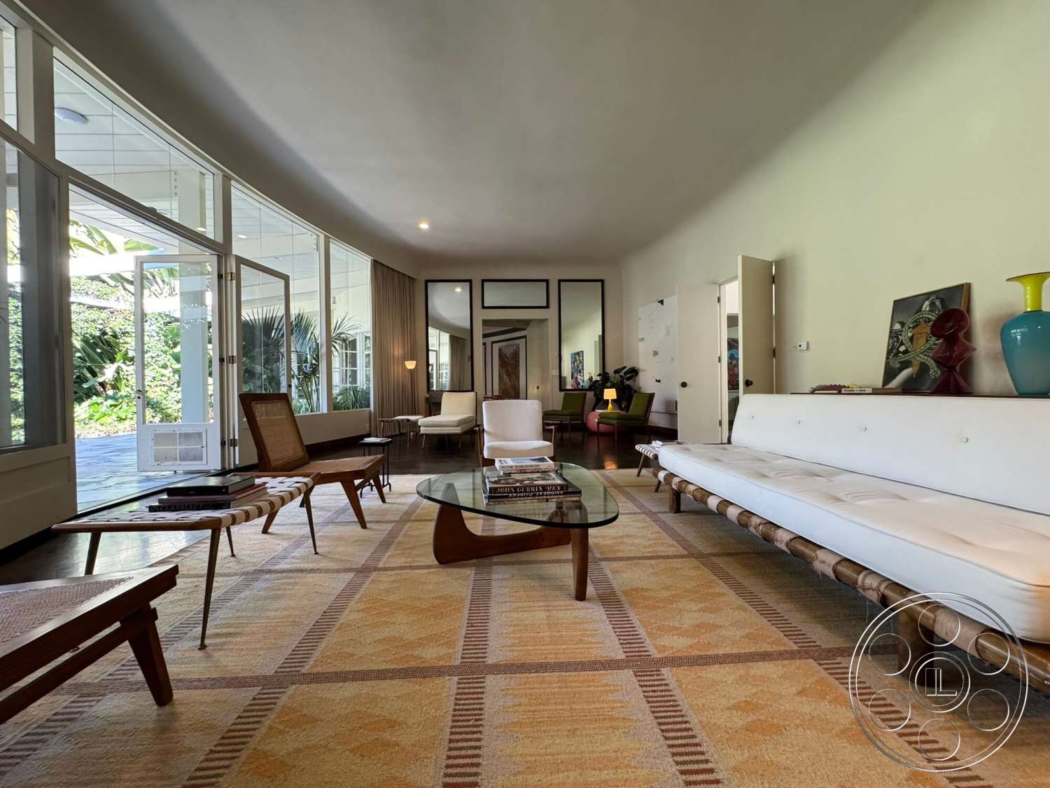 The Hearst House - neutral color palette, textured area rug, wooden coffee table, open floor plan, concrete flooring, decorative art pieces, white upholstered sofa, large floor-to-ceiling windows, glass side tables, wooden accent chairs, potted indoor plants, exposed wood ceiling, mid-century modern interior, earth-tone wall treatments, glass doors leading to outdoor space