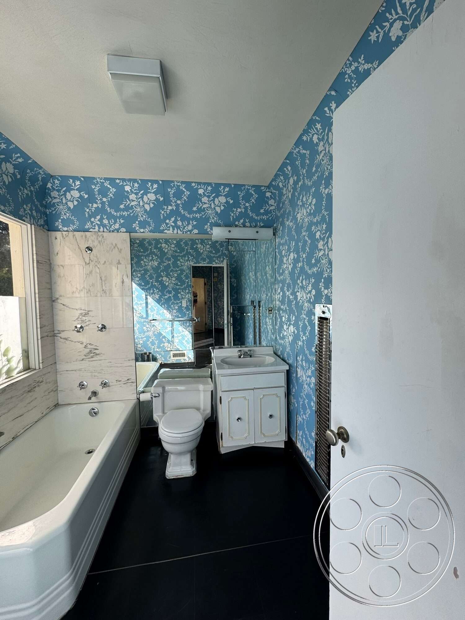 The Hearst House - white porcelain toilet, linear light fixture, sliding glass shower door, interior bathroom, marble bathtub surround, white porcelain bathtub, white vanity with sink, blue floral wallpaper, black tile bathroom flooring, mirror bathroom wall