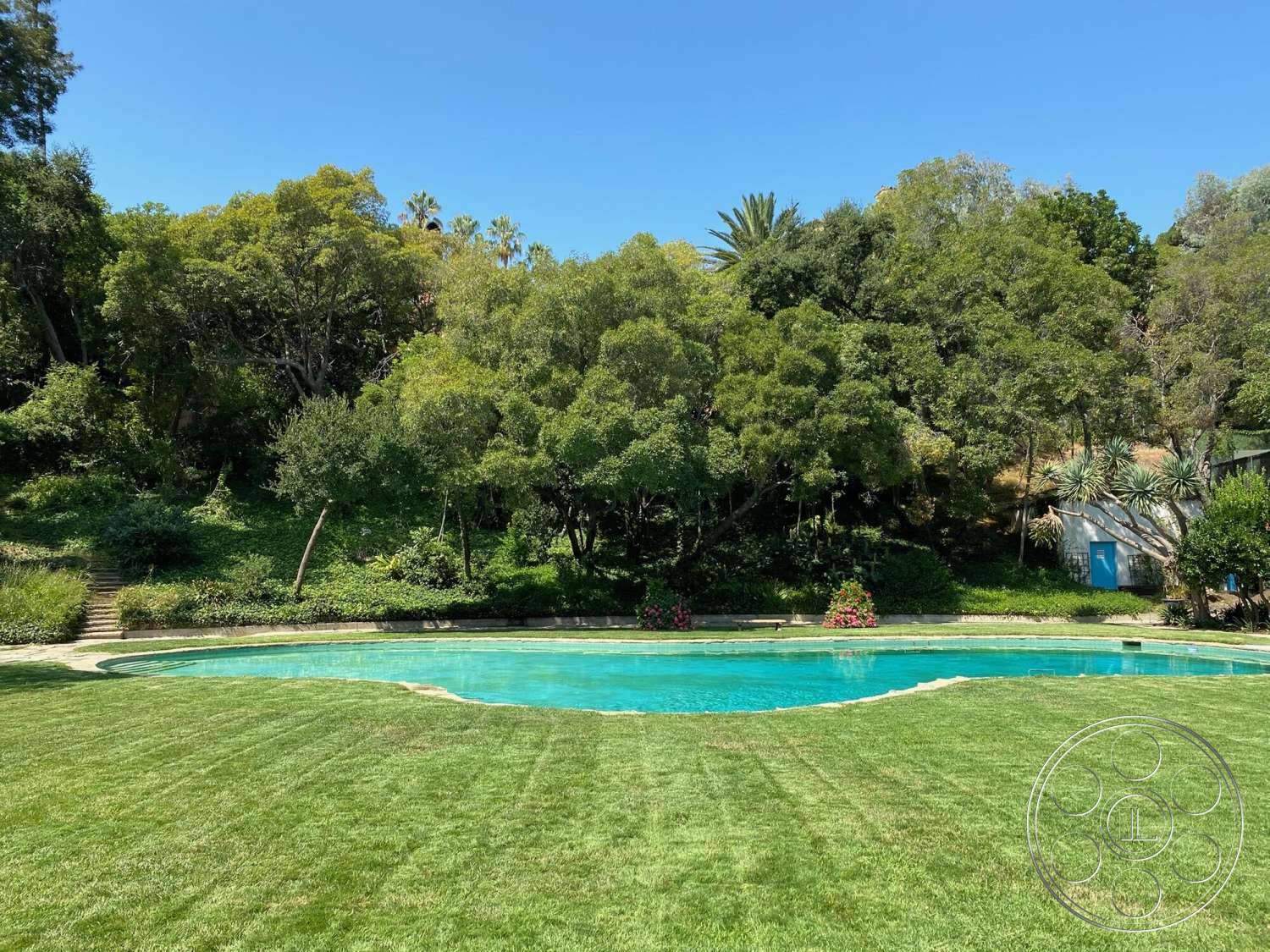 The Hearst House - exterior, landscaped garden, lush green lawn, flowering shrubs, tall trees, blue sky, rectangular pool, concrete pool border