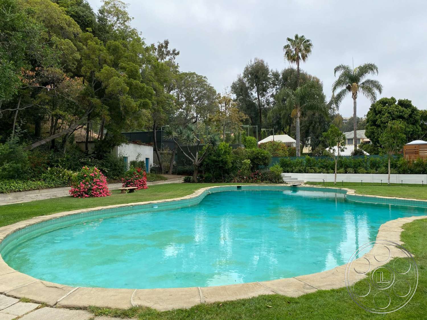 The Hearst House - exterior, lush greenery, tropical palm trees, well-manicured lawn, blue pool water, concrete patio flooring, natural stone pool border, tranquil outdoor setting, kidney-shaped pool, flower garden with pink flowers, surrounding landscaping