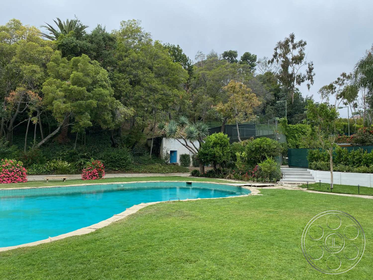 The Hearst House - exterior, landscaped garden, lush greenery, wooden steps, lawn area, vibrant flower beds, stucco wall, concrete pool border, fenced tennis court area, kidney-shaped pool, tropical trees backdrop, blue door accent