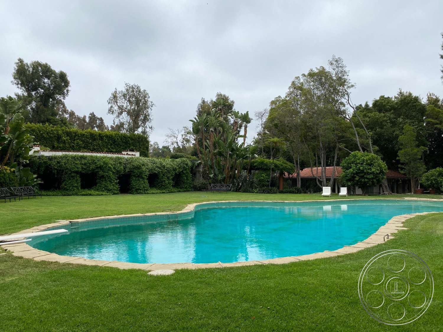 The Hearst House - exterior, lush green lawn, tropical landscaping, hedge privacy screen, oval pool, stone pool border, wooden deck chairs, tree-lined backyard, brick patio roof