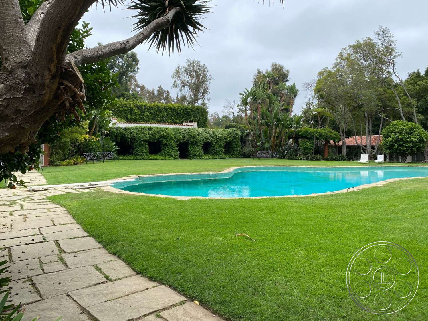 The Hearst House - exterior, patio area, stone pathway, lush green lawn, metal outdoor furniture, tropical landscaping, drought-resistant plants, hedge privacy wall, oval pool, sunny garden setting