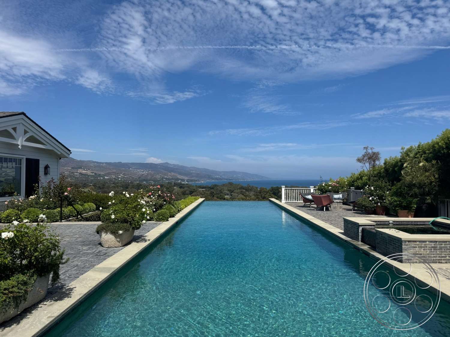 Americana 113 - modern architectural style, infinity pool, outdoor lounge chairs, mountain view setting, stone patio flooring, concrete pool border, lush greenery landscaping, exterior residential home, blue sky with clouds, landscaped garden with flowerbeds, white wooden railing, brick spa surround