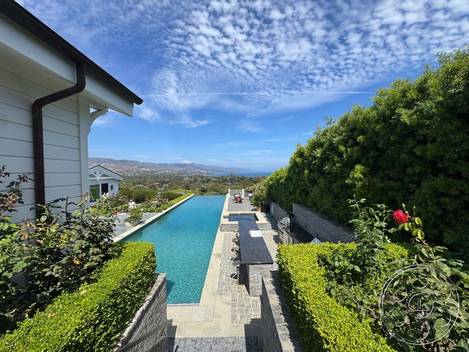 Americana 113 - exterior, landscaped garden, textured stone wall, outdoor dining area, clear blue sky, poolside lounge chairs, infinity pool, rectangular pool, stone patio flooring, concrete pool border, modern outdoor design, surrounding hills view, low-maintenance hedges
